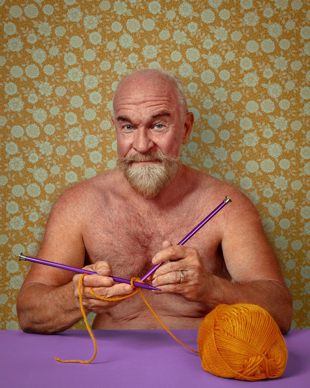 Older male knitting with orange yarn and purple needles against a floral wallpaper background, creating a visually striking composition.